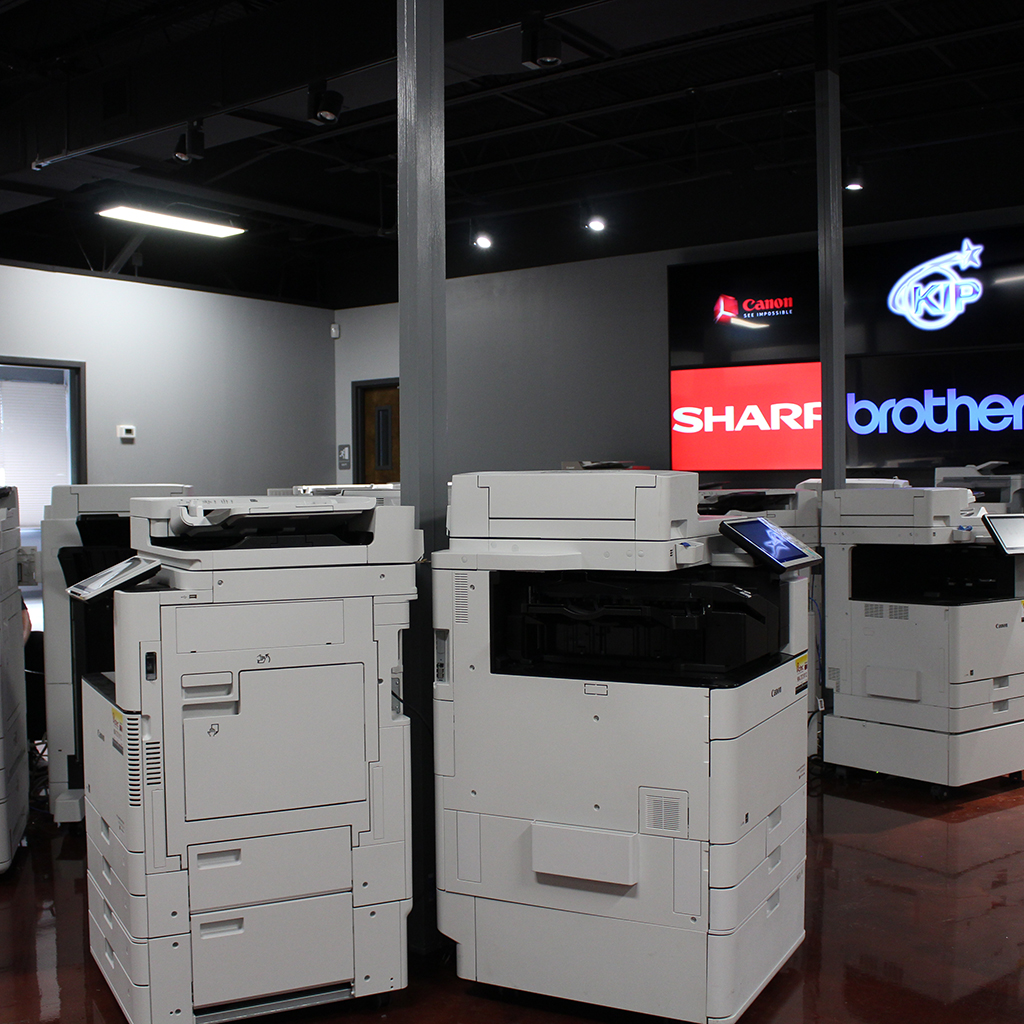 Several multi-function printers are displayed in a showroom. Brand logos including Canon, Sharp, Brother, and Konica Minolta are visible on signs in the background.